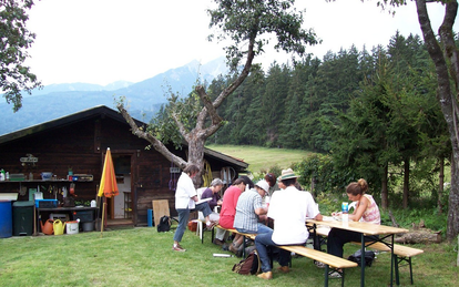 Vor einer Hütte, umgeben von einer Wise und einem Wald im Hintergrund, sitzen mehrere Menschen an einem Tisch und nehmen an einem Kurs teil.