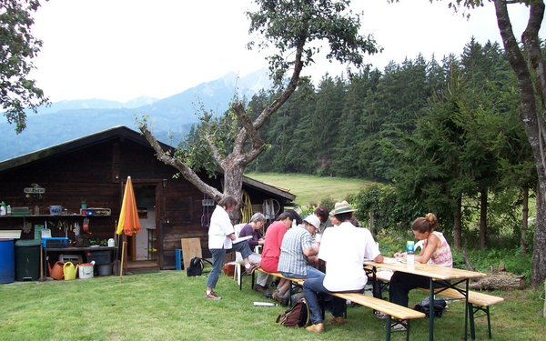Vor einer Hütte, umgeben von einer Wise und einem Wald im Hintergrund, sitzen mehrere Menschen an einem Tisch und nehmen an einem Kurs teil.